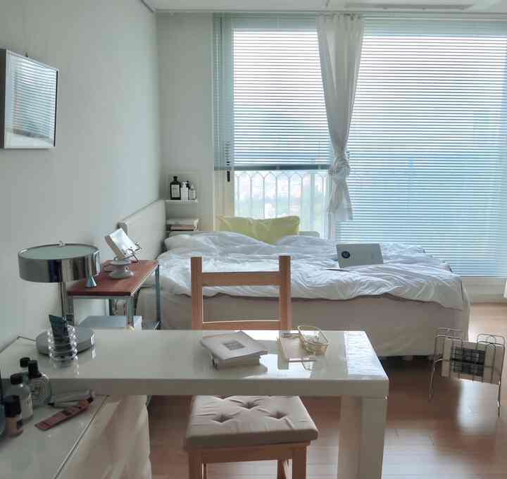 Small bedroom with white and wood tones, featuring a central bed, desk, and blinds on the window creating a clean atmosphere