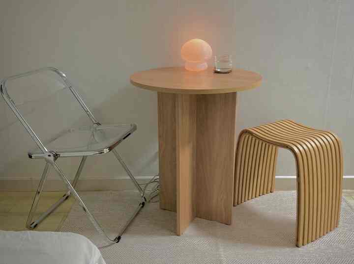 Natural color toned compact studio apartment featuring a round wooden dining table, transparent folding chair, and wooden stool creating a cozy home cafe space