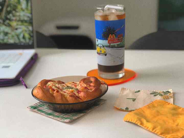 Cozy home cafe setup on a white desk with an orange coaster, blue designed glass, and transparent plate with cake in center