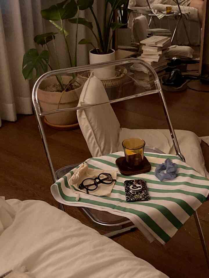 Natural studio bedroom with wood tone flooring and white bedding, featuring a transparent folding chair, rattan mini table, and plants