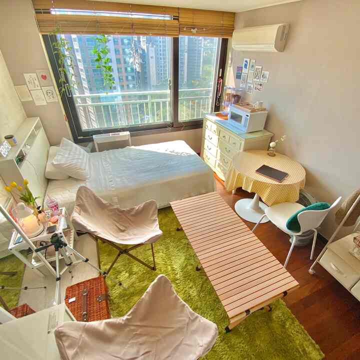 明るい自然光が差し込むナチュラルトーンの狭い寝室、白いベッドと円形のダイニングテーブル、緑のラグ、ハンギングプランツが配置された居心地の良い空間