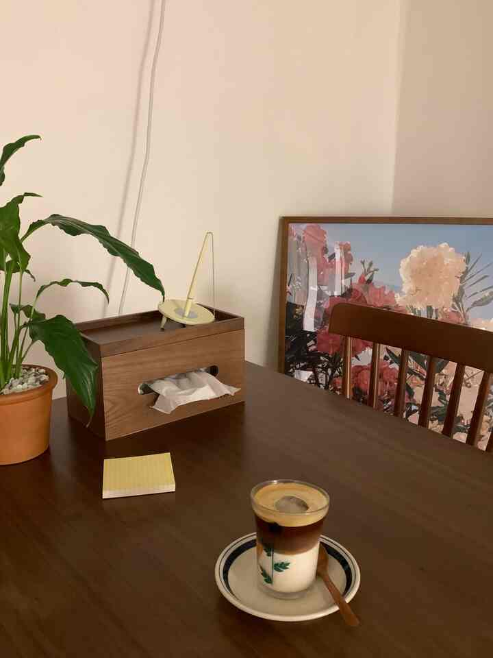 落ち着いたウッドトーンのダイニングルーム、テーブル上にコーヒーと植物があり、背景に花の額縁が配置された雰囲気のある空間