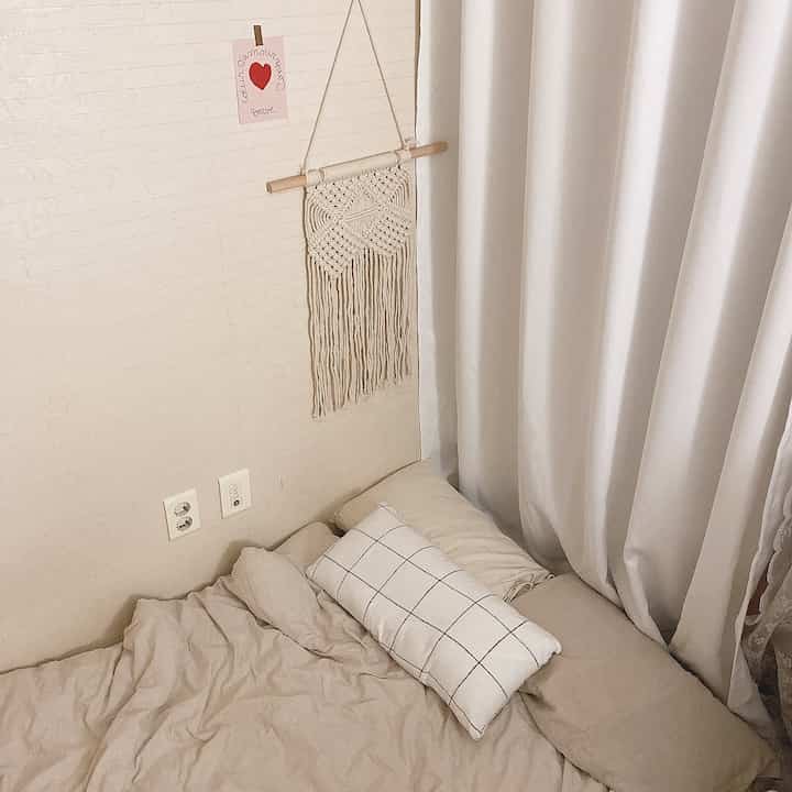 ベージュとアイボリーの色調の寝室、枕とマクラメの壁飾りが配置されたシンプルで温かみのあるインテリア