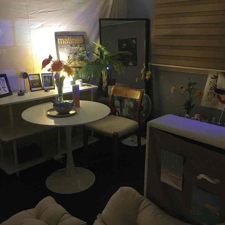 暖かい照明のナチュラルトーン寝室、白い円形ダイニングテーブルと木製の椅子、フロアランプと植物が配置された居心地の良い空間