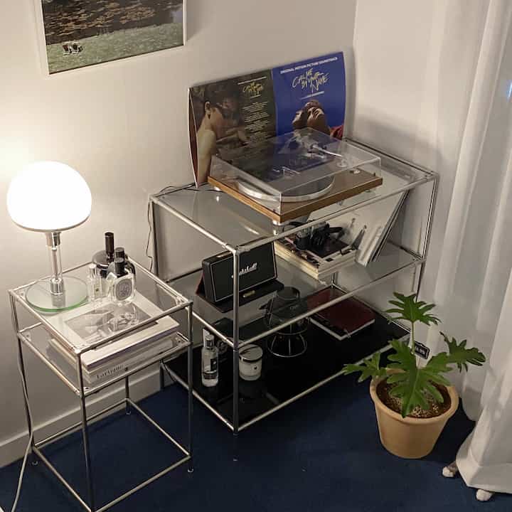 Modern personal room with blue carpet and modular metal shelves featuring turntable, stand lamp, and a potted plant creating cozy atmosphere