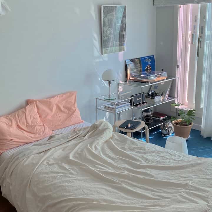 Pastel pink pillows and white bedding in a cozy bedroom with blue carpet flooring, illuminated by natural light in a small room