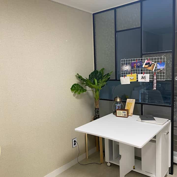 White-toned studio apartment space featuring a folding dining table and large plant creating a cozy atmosphere