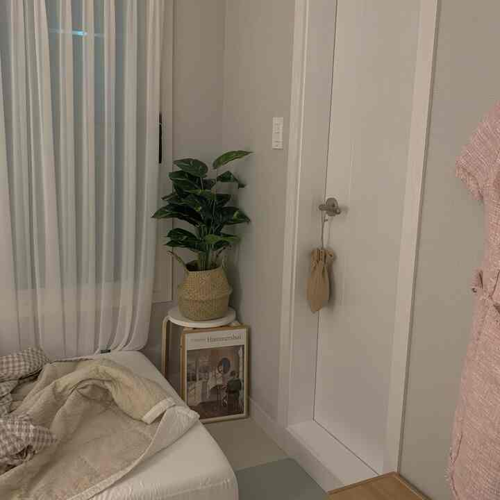 Natural-toned studio bedroom featuring beige bedding, a stool with a green plant, and a framed poster creating a cozy atmosphere