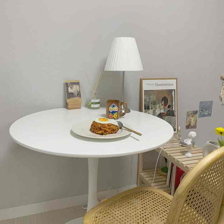 White-toned studio dining room featuring a round dining table, wood-tone chair, and table lamp with a simple atmosphere