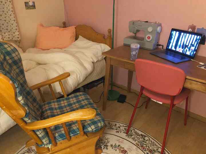 A small room with pink walls and natural wood-tone furniture featuring a bed, rocking chair, and laptop on desk creating a cozy relaxation space.