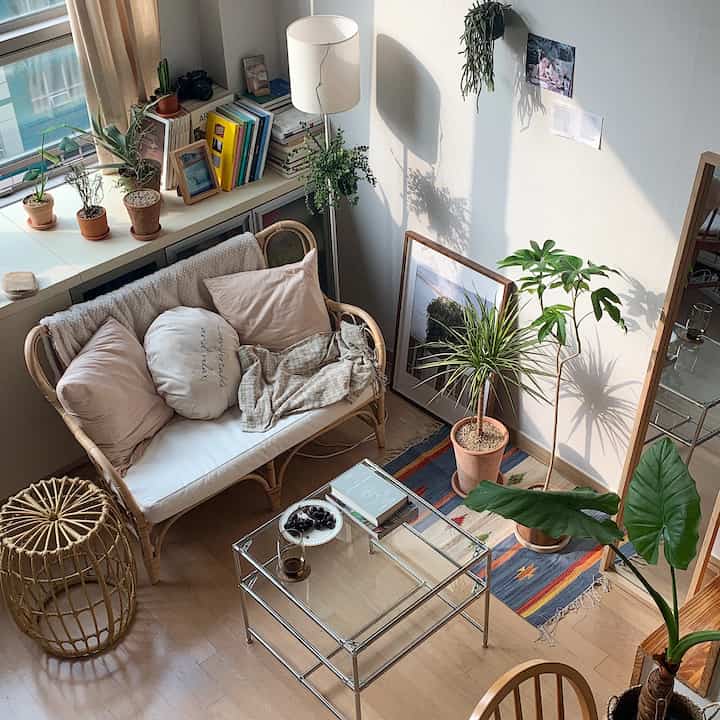 Natural-toned compact living room featuring rattan sofa, glass table, various plants, and picture frames in a cozy setting