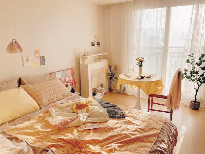 Warm beige and yellow-toned bedroom featuring a cozy bed, lace curtains, and a round table with a yellow checkered tablecloth creating a comfortable atmosphere