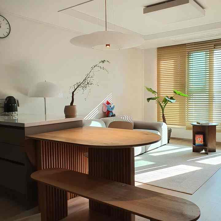 A white and wood-toned living room featuring a round dining table, sofa, blinds, and plants creating a warm atmosphere