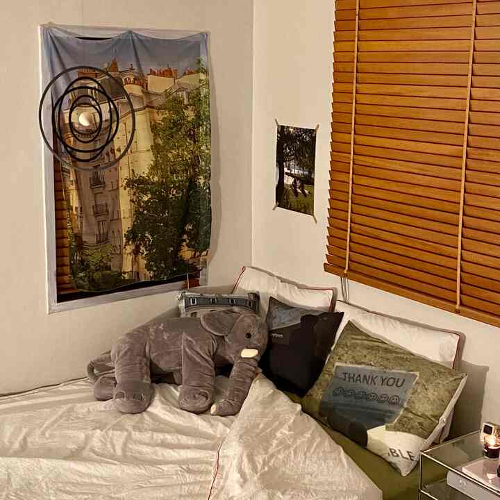 Cozy bedroom featuring wood tone blinds and white bedding, decorated with an elephant plush and a mobile for a comfortable vibe