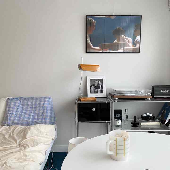 Modern bedroom with white walls and a round table, featuring a blue check bedspread, turntable, and a lamp on the shelf