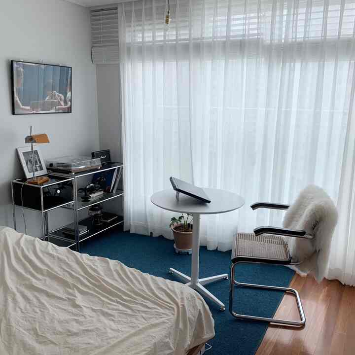 Bright and modern bedroom with white walls and curtains, blue rug, round table, and cantilever armchair