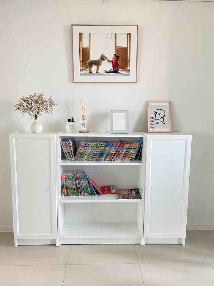 ホワイトトーンのリビングルーム、中央にイケアのBILLY本棚が配置され、上にインテリアフレームとディフューザーが置かれた、清潔感のあるインテリア