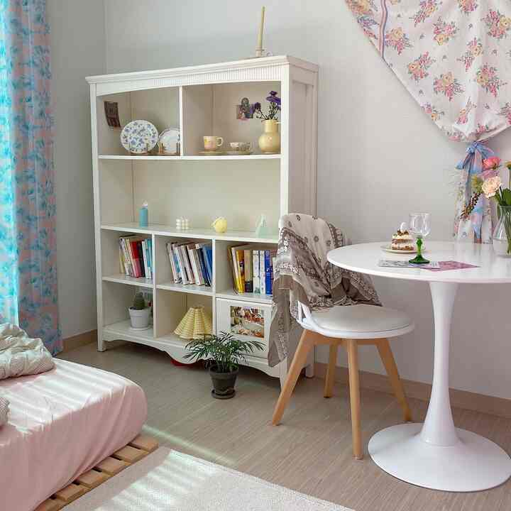 白と木目調が調和した6畳のワンルーム、円形テーブルとダイニングチェア、フローラルカーテン、ベッドと収納棚が配置された雰囲気のあるおうちカフェ空間