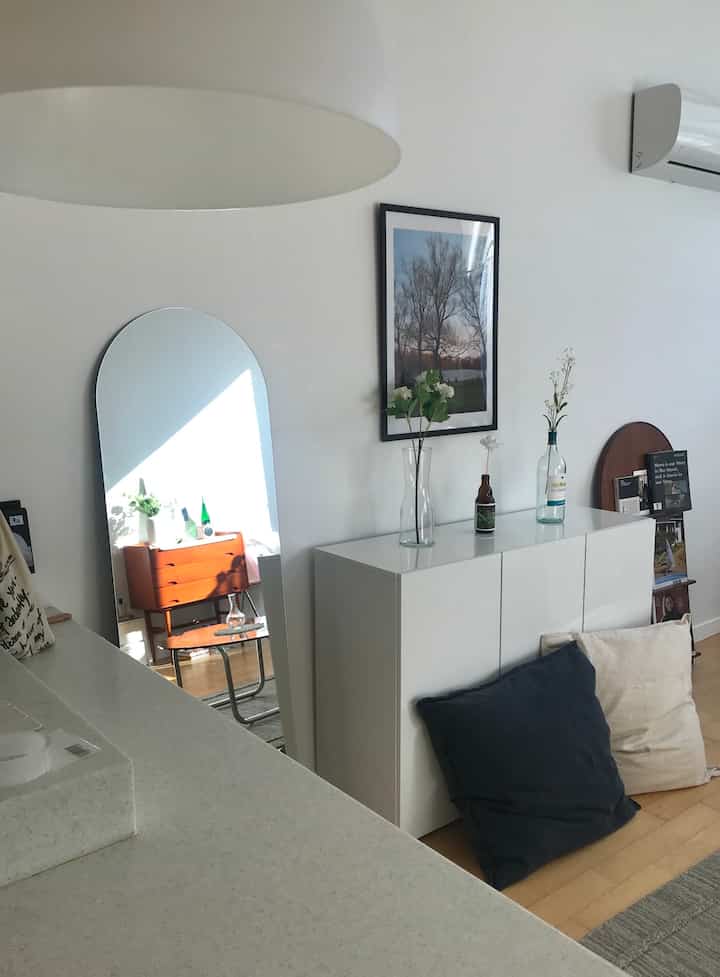 白い壁と木目調の床が調和するリビング、アーチ型の鏡とクッションが配置されたミニマルでナチュラルな雰囲気のインテリア