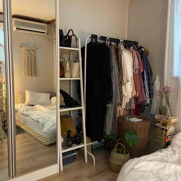 白とナチュラルカラーのワンルーム寝室、ラックやラタンバスケット、植物が配置されたシンプルな空間