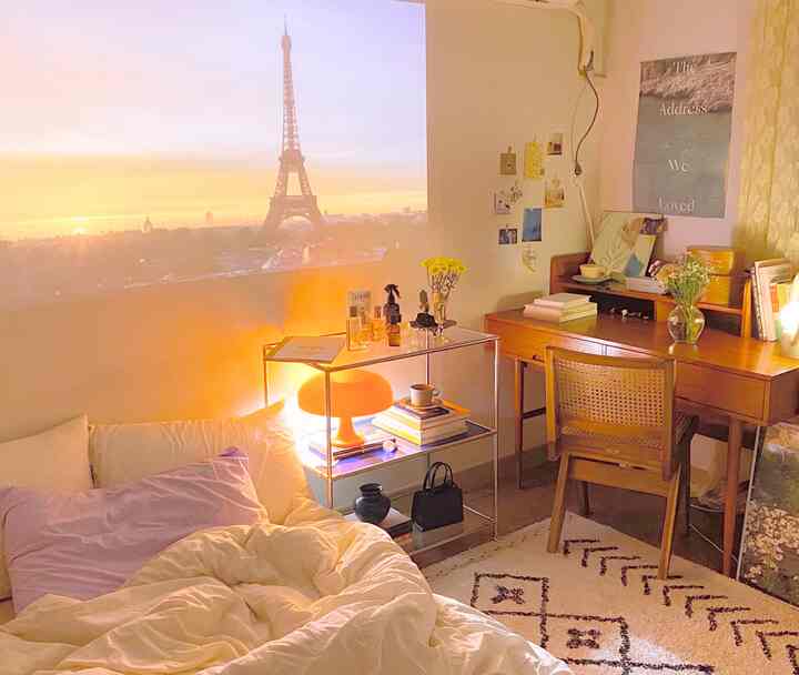 Warm-toned studio bedroom featuring beige and lavender bedding, wooden desk, and an orange lamp creating cozy interior ambiance