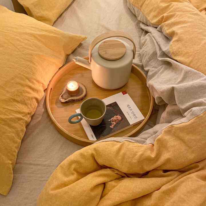 暖かい黄色とベージュのトーンが調和する寝室のベッド上に、リネン寝具と木製トレイが置かれ、陶器の急須、本、キャンドルが配置された雰囲気のある空間