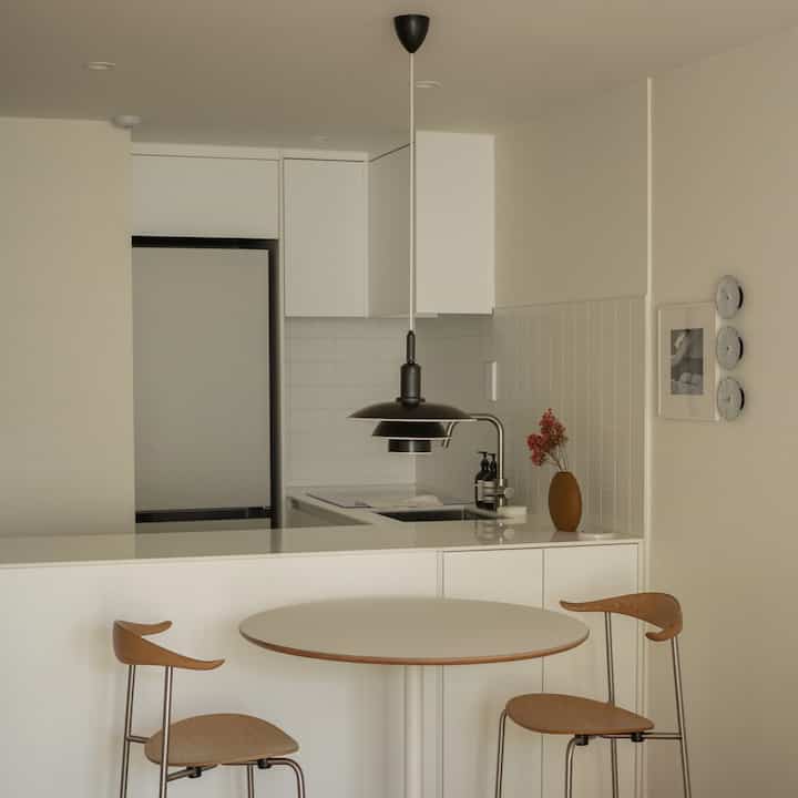 ホワイトと木目調の調和が美しい、コンパクトな2人用キッチン・ダイニングルーム。モダンなペンダントライトが配置された清潔感のある空間