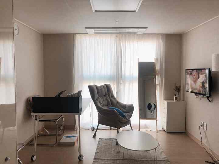 Beige and white toned living room for single household, featuring transparent curtains, armchair, desk, and wall-mounted TV in a simple modern setting
