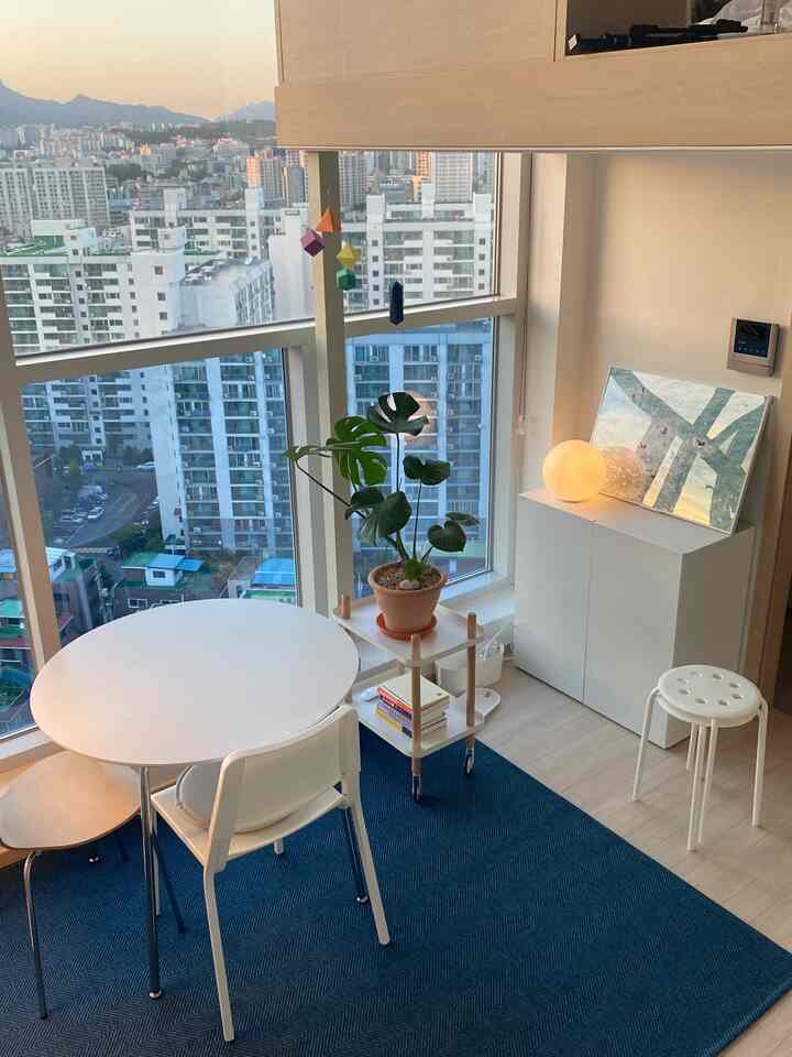 A white and blue toned apartment home office featuring a round dining table, chairs, large windows with city view, natural light, and a green plant