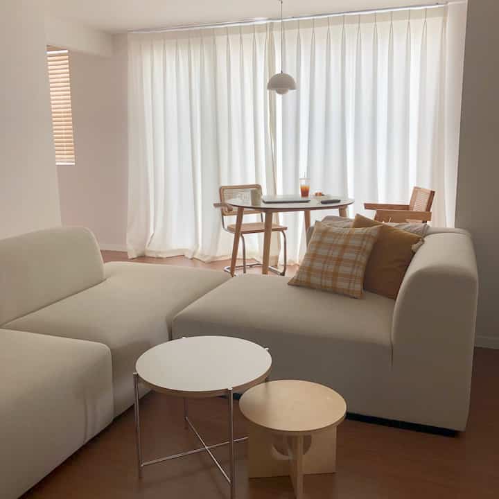 柔らかいホワイトとウッドトーンの雰囲気あるリビング、快適なソファとラタンチェア、小さなテーブルとペンダントライトが配置された印象的な空間