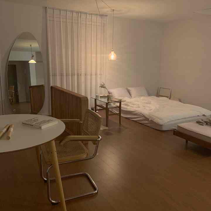 A natural modern bedroom with white bedding and wood tones, featuring a mattress, armchair, and table in a minimalist setting