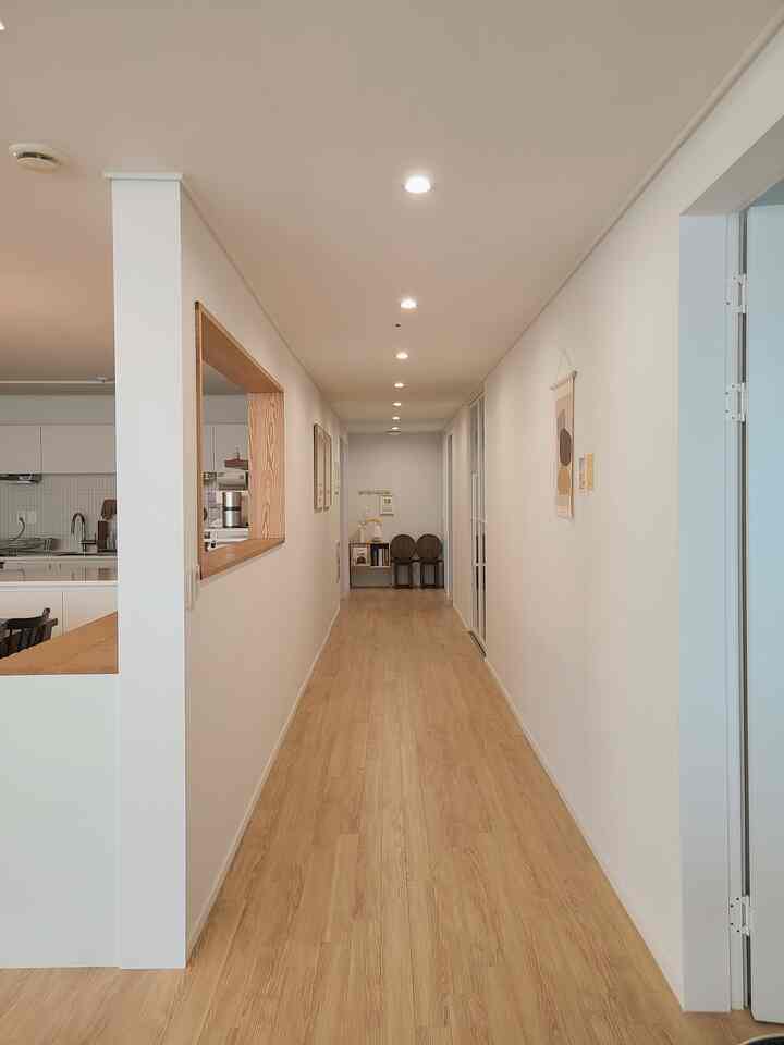 Natural and minimal hallway with wood tone flooring, white walls, and kitchen connected through a window opening