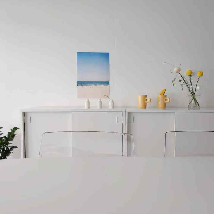 White-toned living room featuring transparent chairs and simple table with yellow mugs and vases, creating a clean and modern atmosphere