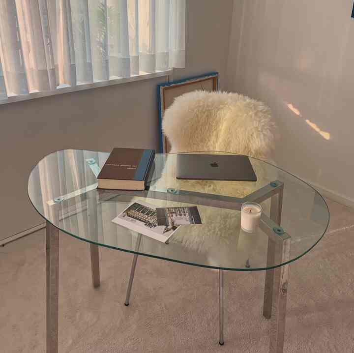 A small home office in soft white and natural tones, featuring a glass table and fluffy chair creating a cozy atmosphere