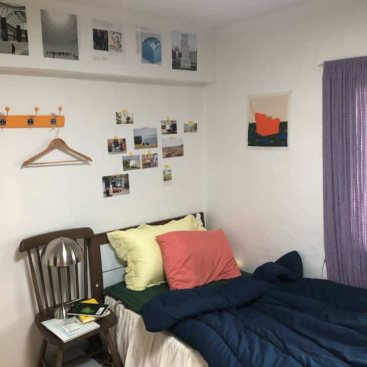 白い壁と紫色のカーテンがある寝室、木製の椅子と布製ポスターが配置された雰囲気のあるヴィンテージインテリア