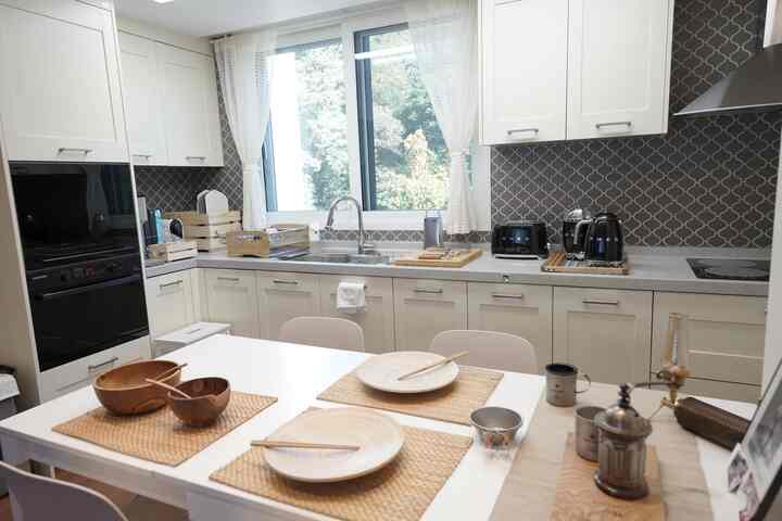 A clean kitchen in white and natural tones featuring a large window, dining table, and black SMEG kitchen appliances