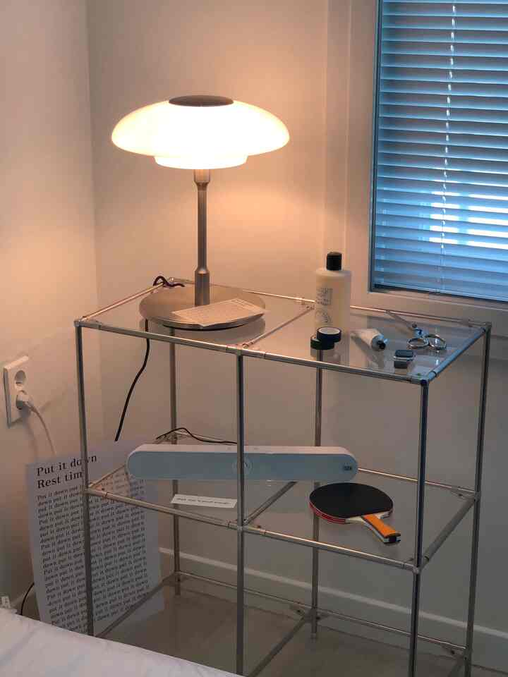 Simple bedroom space featuring a modular metal rack nightstand with glass shelves and a warm glowing table lamp
