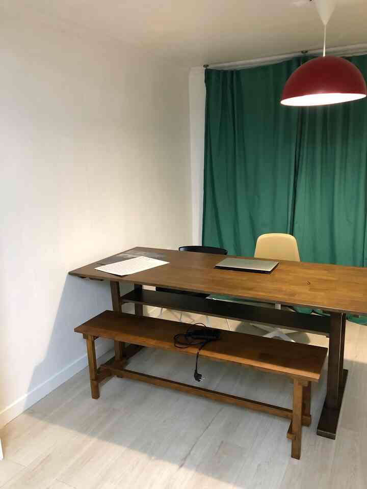 Natural tone workspace featuring a wooden dining table, bench, green curtains, and a red pendant light creating a cozy atmosphere
