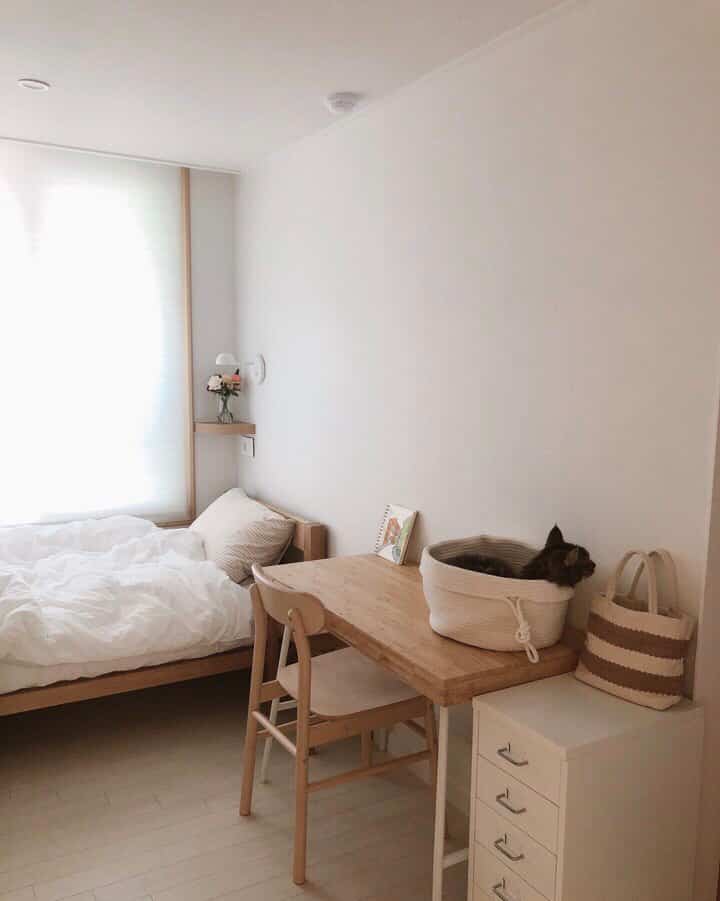 White and wood-tone bedroom featuring minimal furniture with a cat resting in a cotton basket on the desk, cozy atmosphere