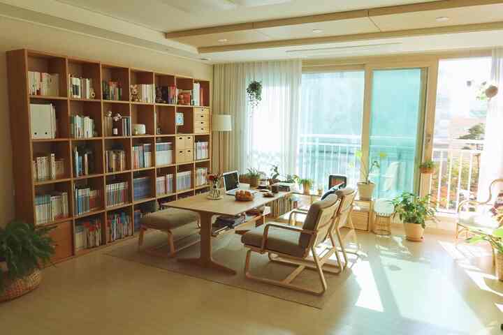 Bright natural-tone living room featuring wooden table, armchairs, bookshelf, curtains, and various plants creating cozy atmosphere