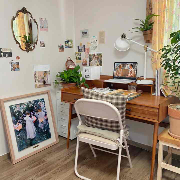 Natural wood tones and white walls define this study space featuring a wooden desk, folding chair, multiple plants, and a vintage mirror creating a cozy work area
