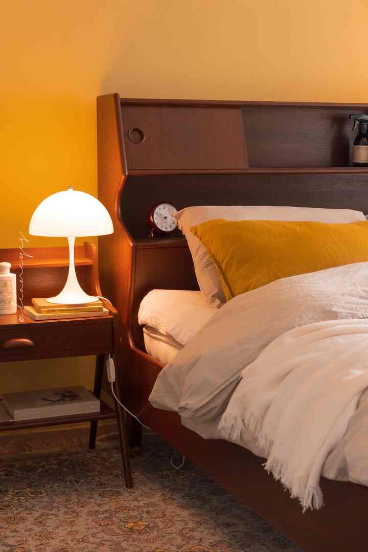 Cozy winter bedroom featuring warm yellow walls, mid-century modern wooden bed and nightstand, with a white table lamp illuminating the space