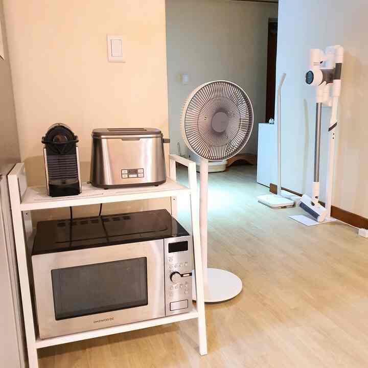 白とベージュのトーンのキッチン空間、金属製棚に電子レンジ、トースター、カプセルコーヒーマシンが置かれ、無線扇風機とコードレス掃除機が配置された清潔なインテリア