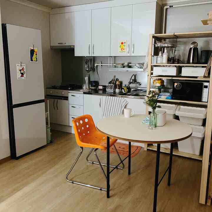Compact kitchen with white and natural tones, featuring a round dining table and orange chair in a tidy space