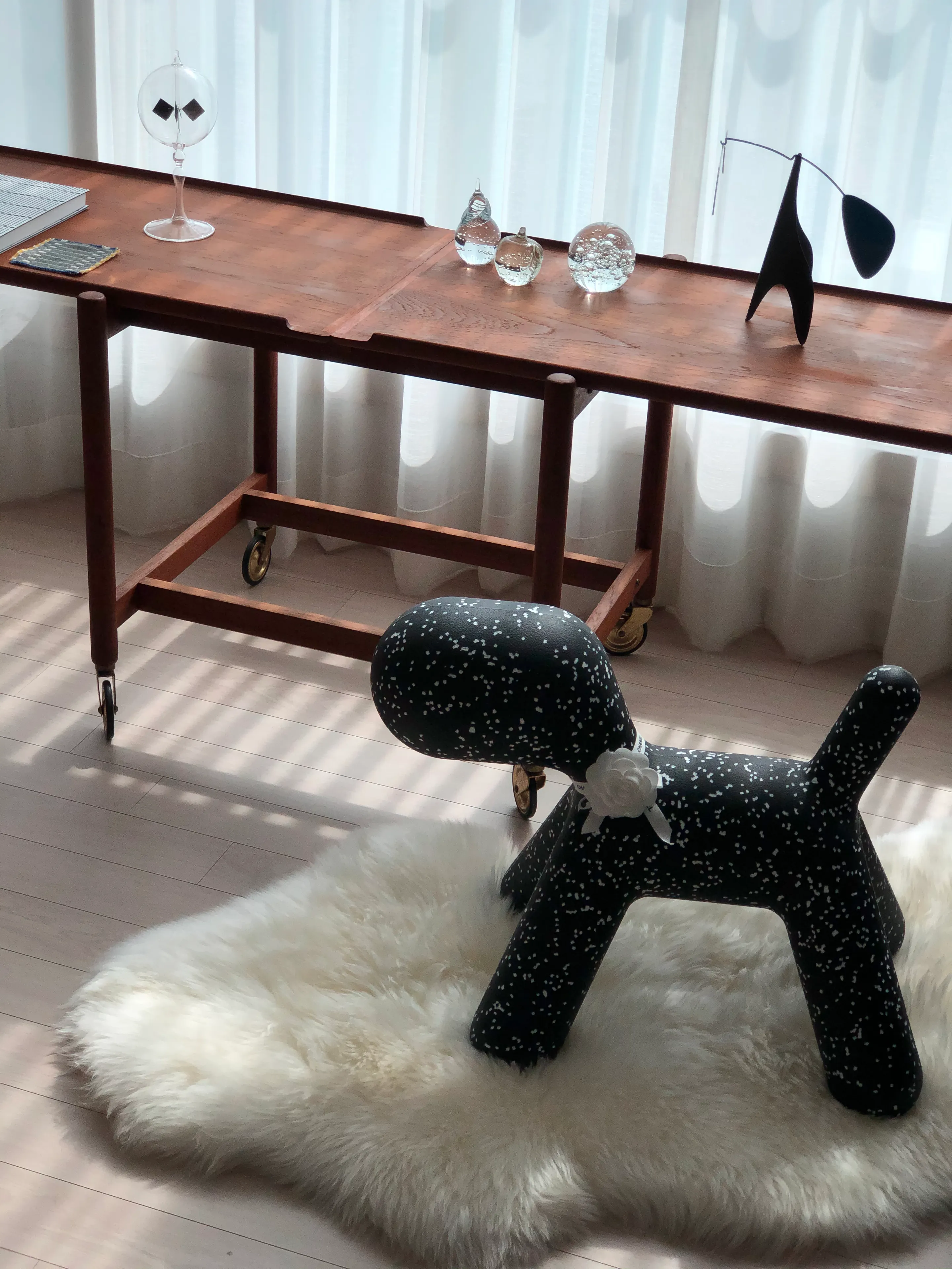白いカーテンの前に木製のテーブル、白い羊毛ラグの上に黒い犬の形のオブジェが配置された温かみのある木目調のリビングスペース