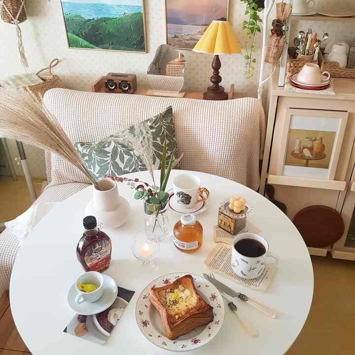 ベージュとナチュラルカラーのワンルームリビング、丸いダイニングテーブルとクッション付きソファ、テーブルランプと額縁が配置された居心地の良いおうちカフェの空間