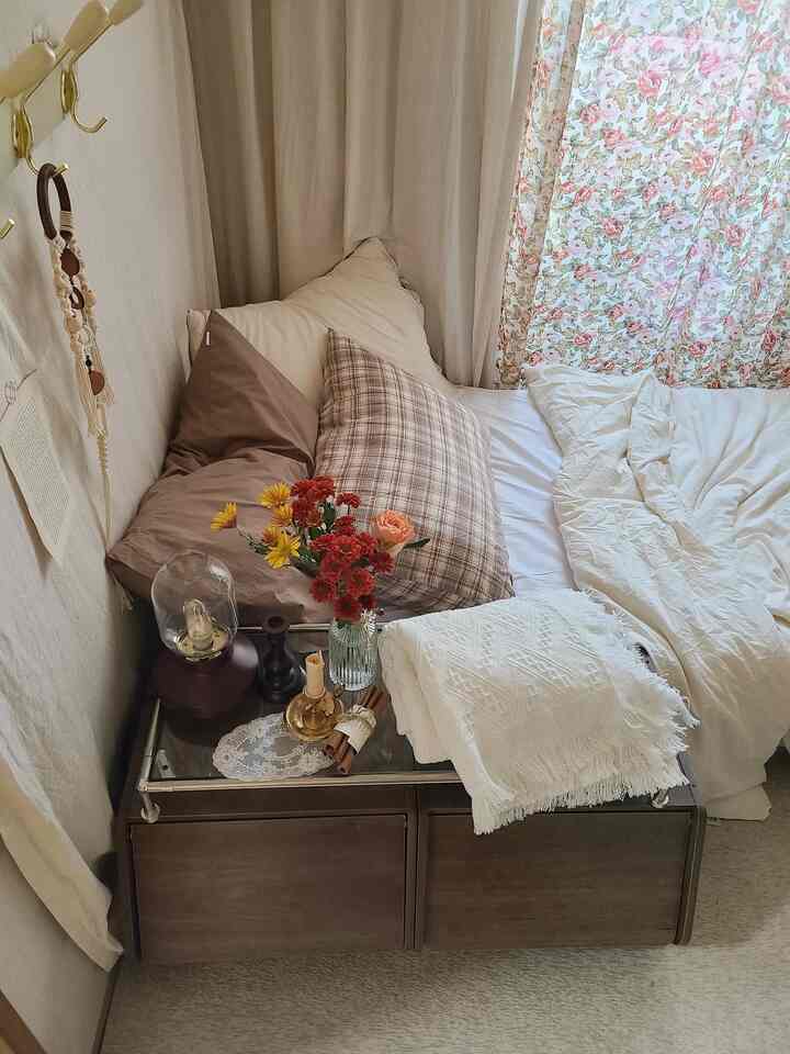ナチュラルなベージュトーンの小さな寝室、ナイトテーブルや多様な枕、花柄カーテンが温かみのある雰囲気を演出する空間