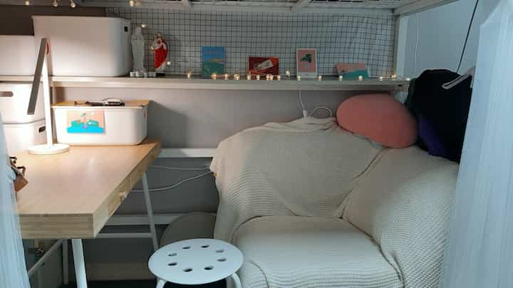 ホワイトとナチュラルトーンのリビング、小さな木製デスクとブランケットが掛けられたソファ、白いスツールが配置された雰囲気のあるコンパクトな空間