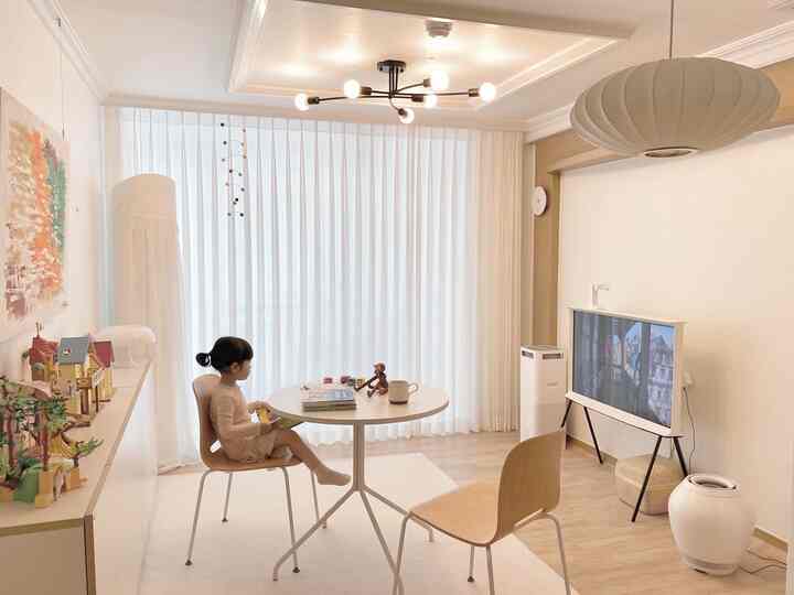 Bright and natural white-toned living room featuring a round table with chairs, a child sitting, curtains, and a mobile in a simple setting