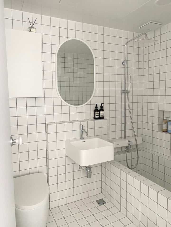 A simple modern bathroom featuring white square tiles on walls and floor, a matte sink, and an oval mirror